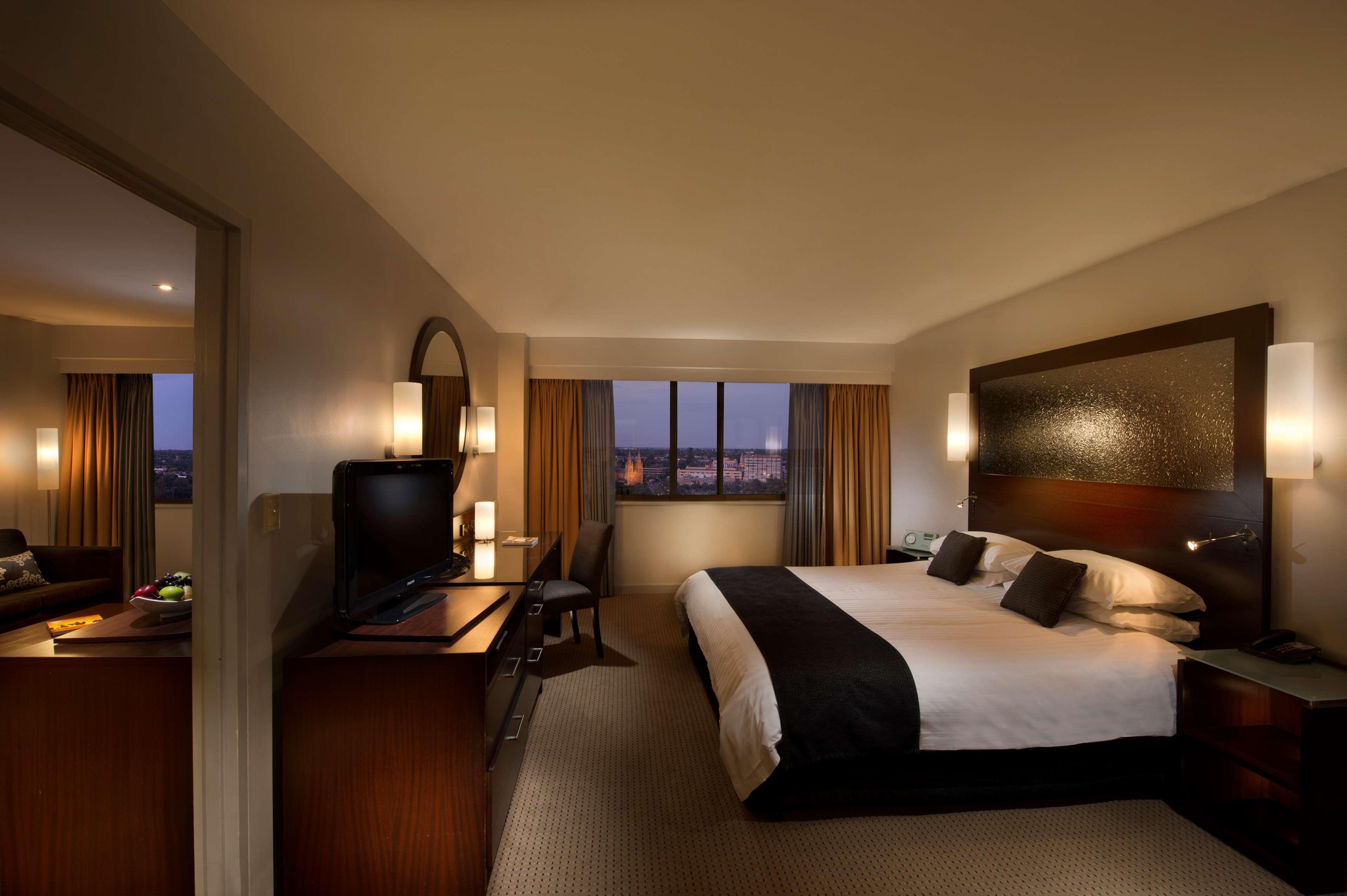 Hotel Stamford Plaza Adelaïde Extérieur photo The photo features a modern hotel room that includes a large bed with dark and light bedding, complemented by two pillows. The room is well-lit with warm lighting and has a contemporary aesthetic. To one side, there is a wooden dresser with a flat-sc