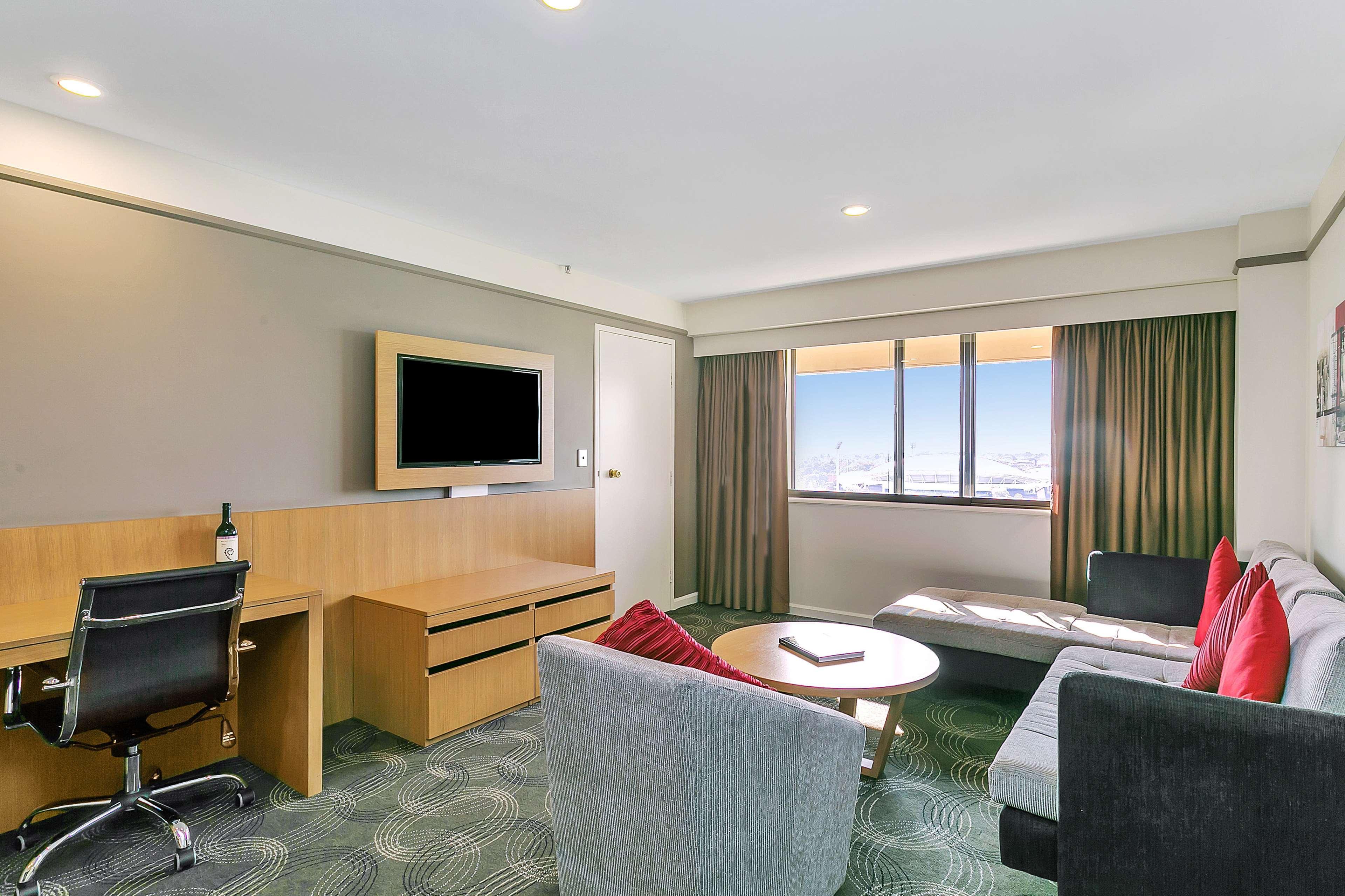 Hotel Stamford Plaza Adelaïde Extérieur photo The photo shows a modern hotel room or suite that features a cozy seating area. There is a comfortable sofa with red accent pillows, a round coffee table in the center, and a sleek desk with an office chair. On the wall, there is a flat-screen TV mou