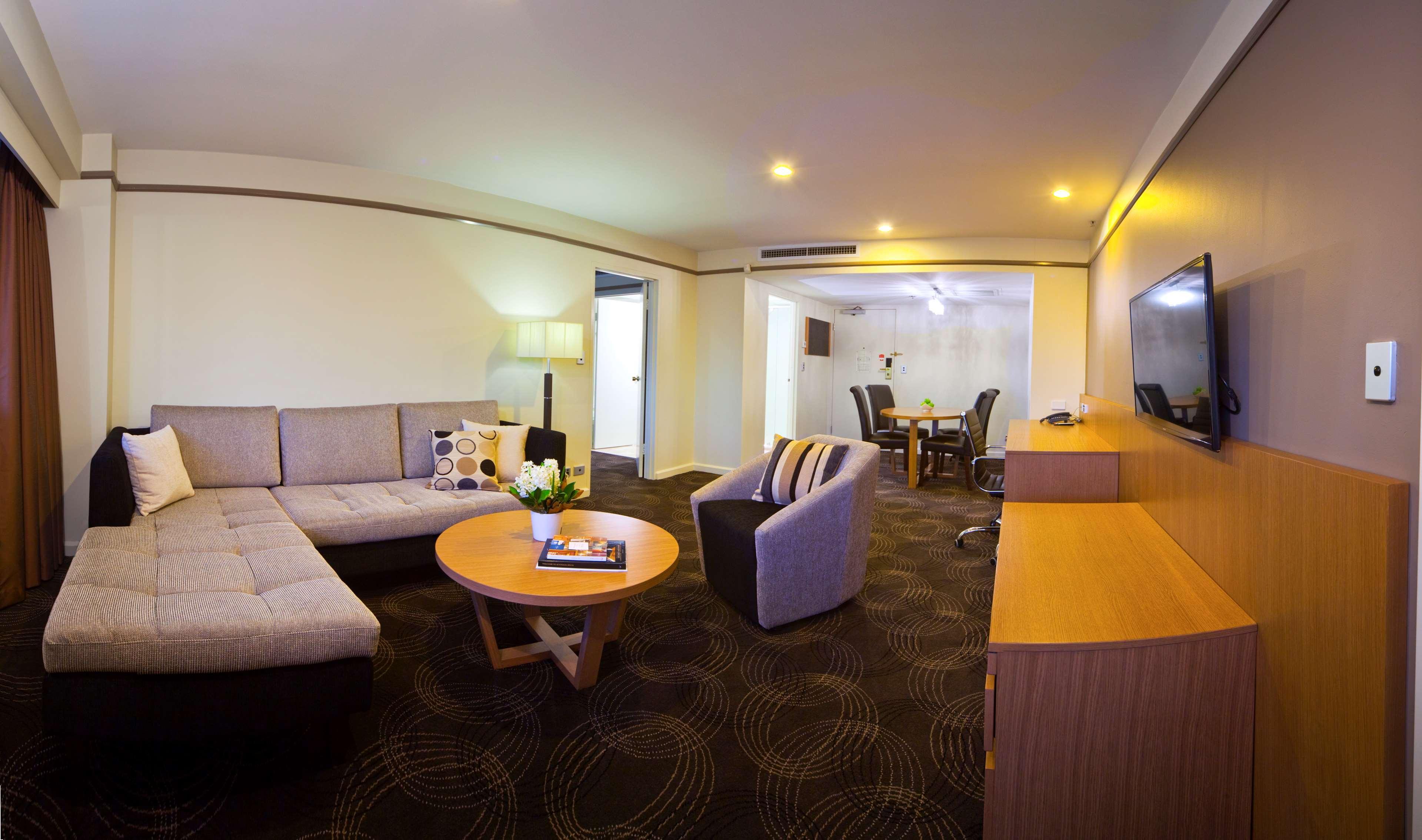 Hotel Stamford Plaza Adelaïde Extérieur photo The photo shows a spacious living area with modern decor. On the left, there is a light gray sectional sofa, accompanied by a round wooden coffee table in front of it. A couple of decorative pillows add a touch of color to the sofa. To the right, the