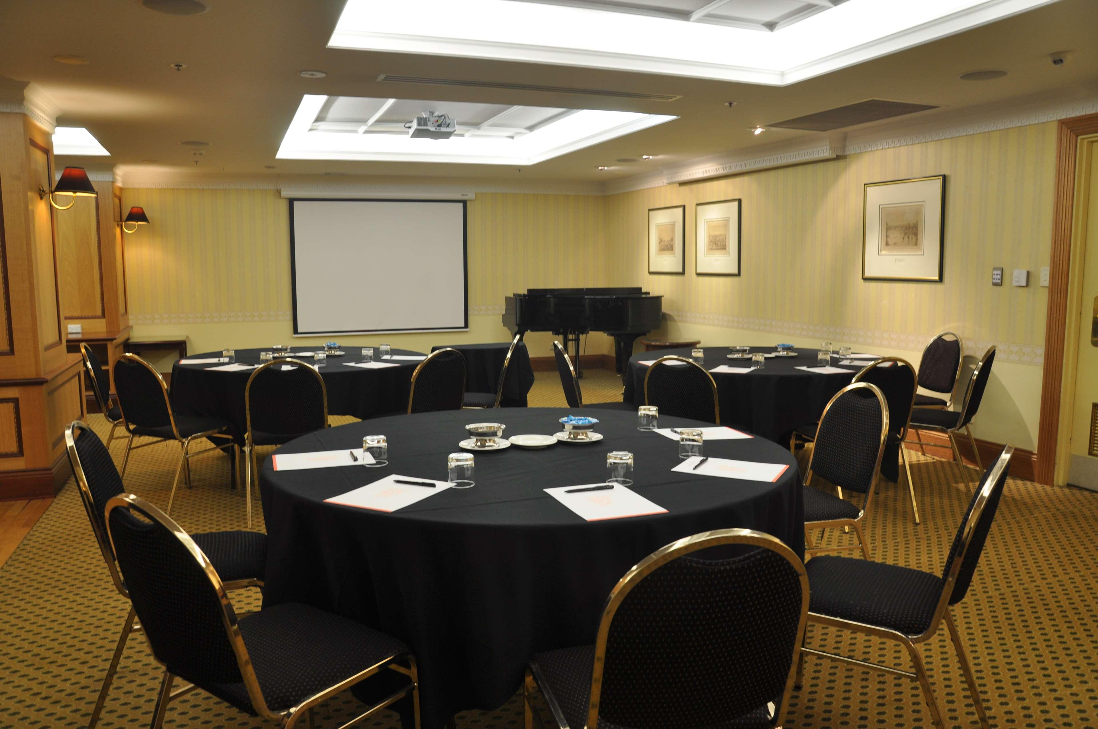 Hotel Stamford Plaza Adelaïde Extérieur photo The photo shows a conference room set up for a meeting or presentation. There are several round tables covered with black tablecloths, each surrounded by black upholstered chairs. On the tables, there are notepads and glasses of water, indicating tha