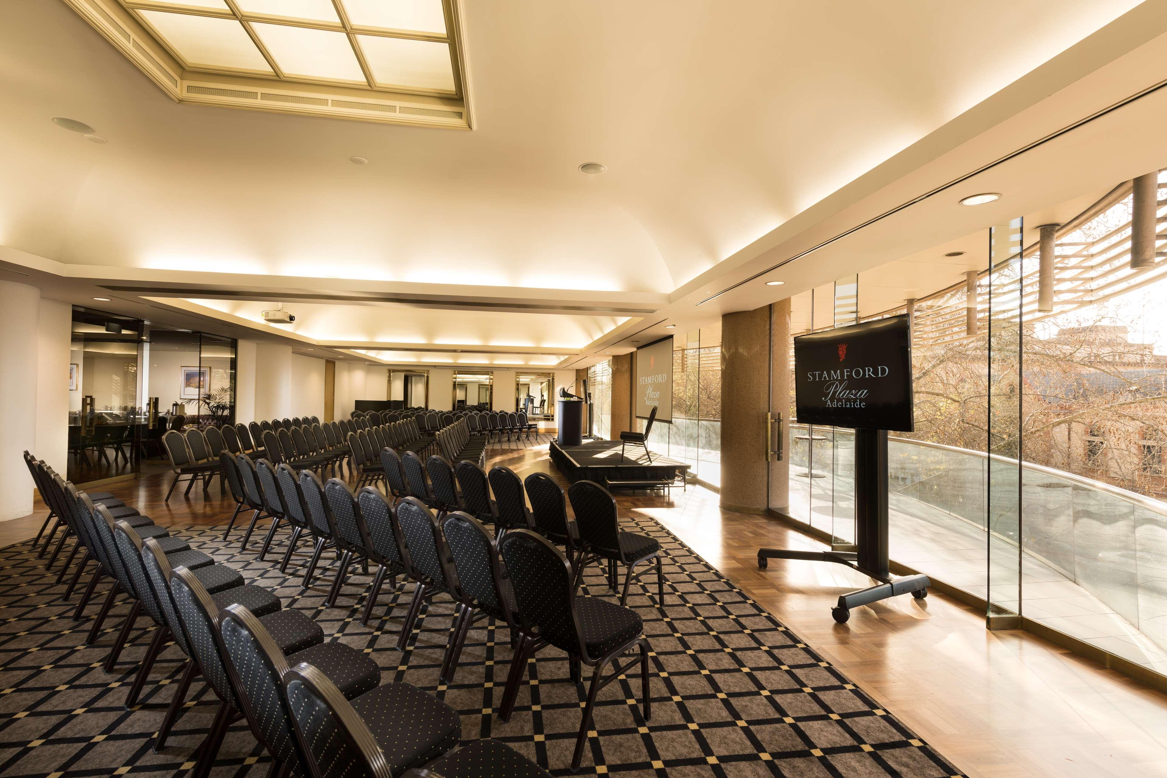 Hotel Stamford Plaza Adelaïde Extérieur photo The photo shows a large, elegantly designed event space or meeting room. There are rows of black chairs set up facing a presentation area. The flooring features a patterned carpet, and large windows allow natural light to enter the room. On one side,
