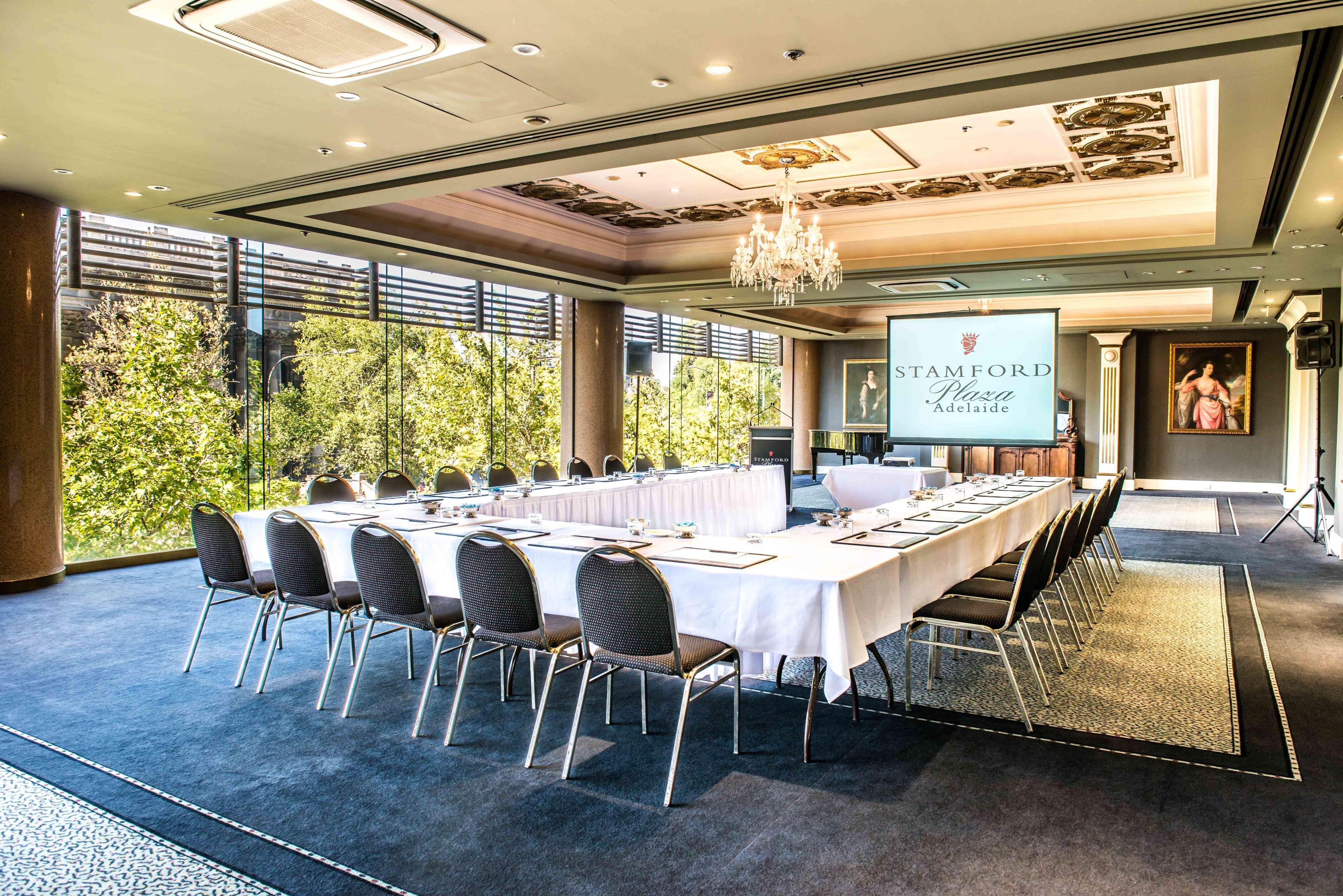 Hotel Stamford Plaza Adelaïde Extérieur photo The photo shows a stylish conference room set up for a meeting or event. There is a long rectangular table covered with a white tablecloth, surrounded by black chairs. A large screen displays the name "Stamford" along with a logo. The room features l