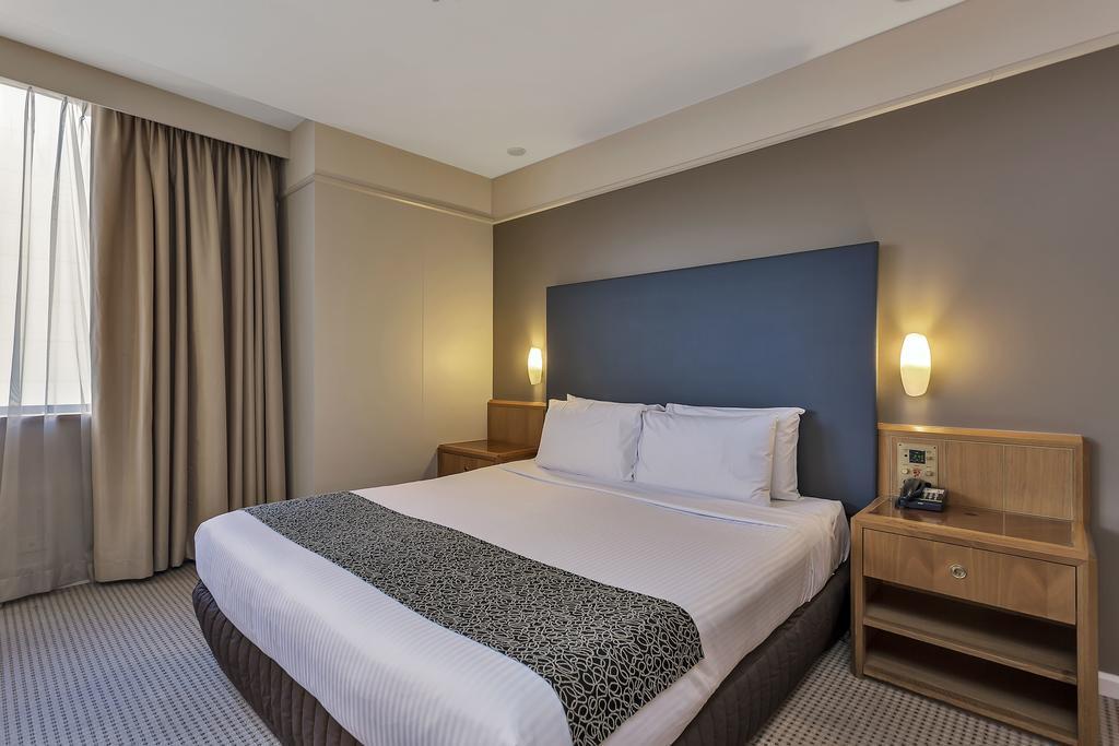 Hotel Stamford Plaza Adelaïde Extérieur photo The photo shows a modern hotel room. It features a double bed with a white duvet and a decorative grey and black throw. The headboard is a solid dark color. There are two bedside tables on either side of the bed, each with a lamp. Natural light enter